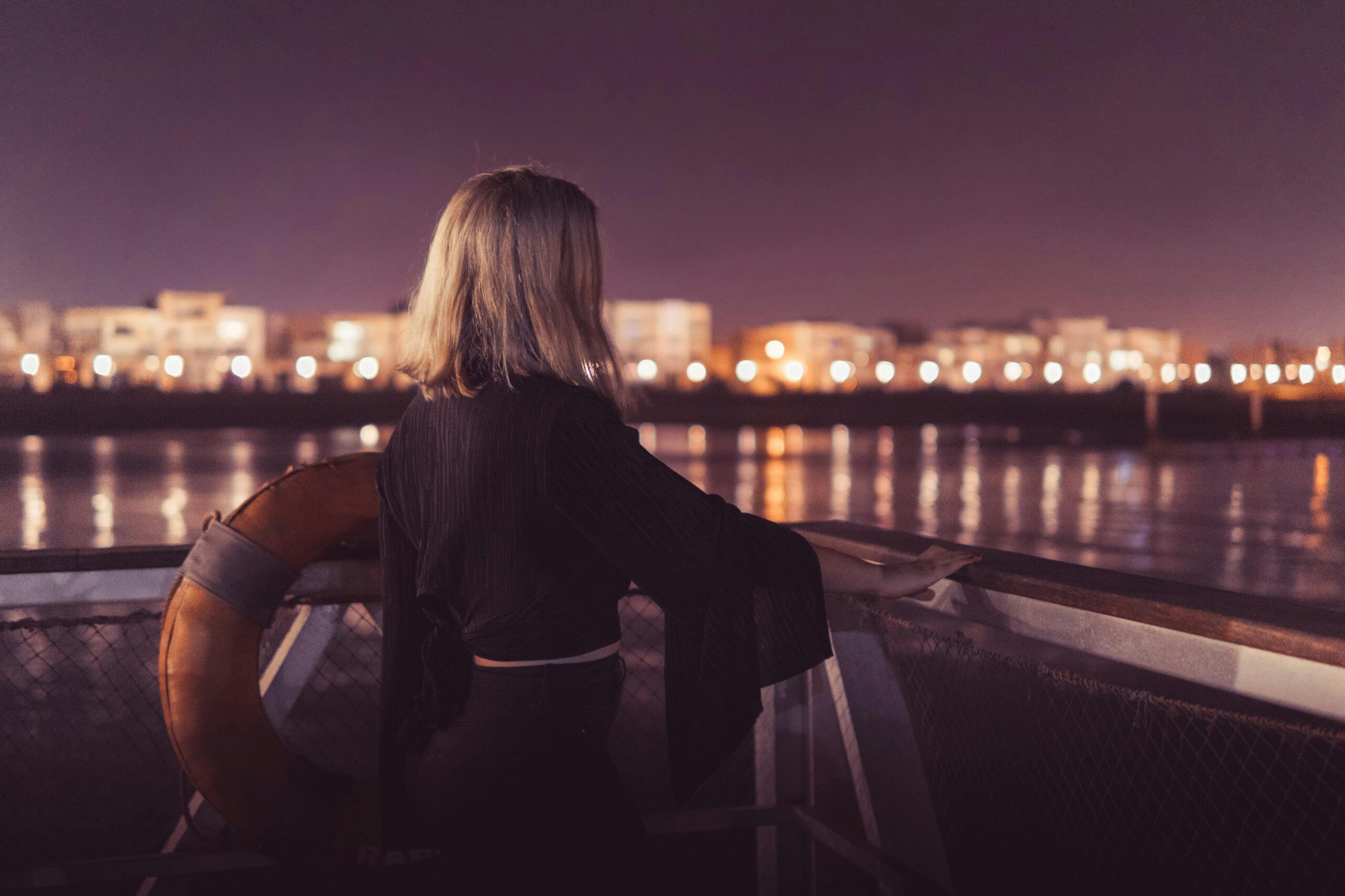 Blonde woman overlooks a lit cityscape across the river at night, evoking calm reflections.